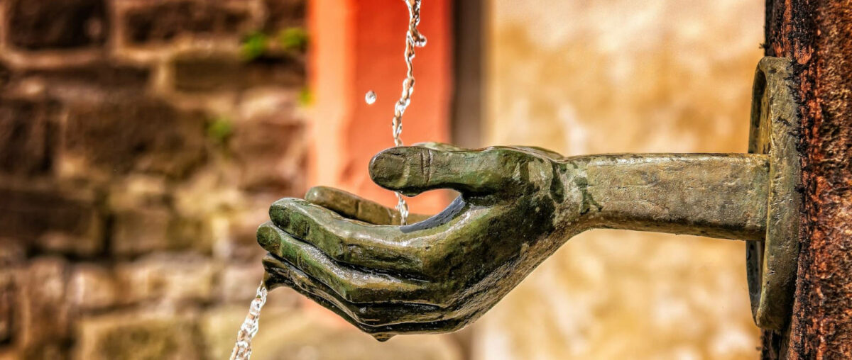 In Skulptur-Hände, welche an einer Säule befestigt sind, fliesst Wasser